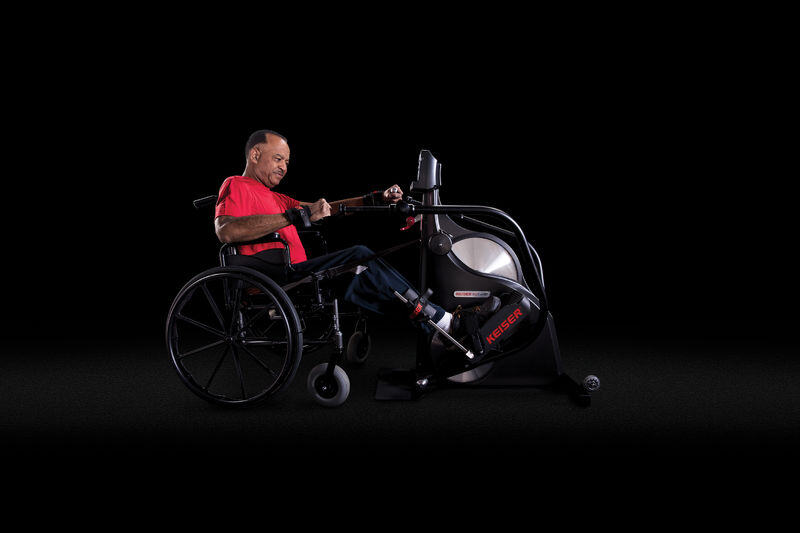 Man in wheelchair operating a Keiser Woman in a assistive scooter pulled up to a Keiser M7i Total Body Wheelchair Accessible Stepper